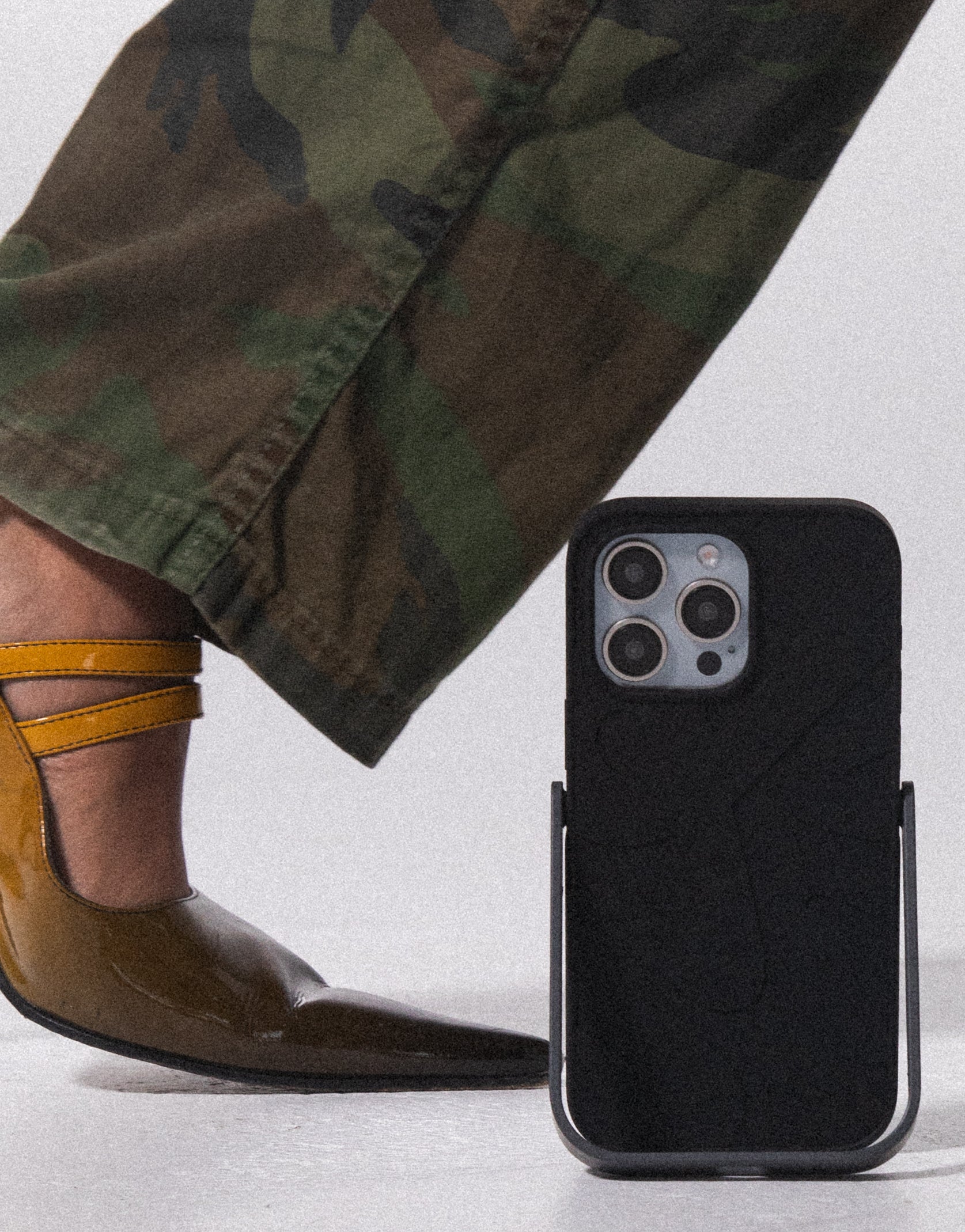 PROPS phone case next to a person wearing camouflage pants and brown shoes on a light gray background