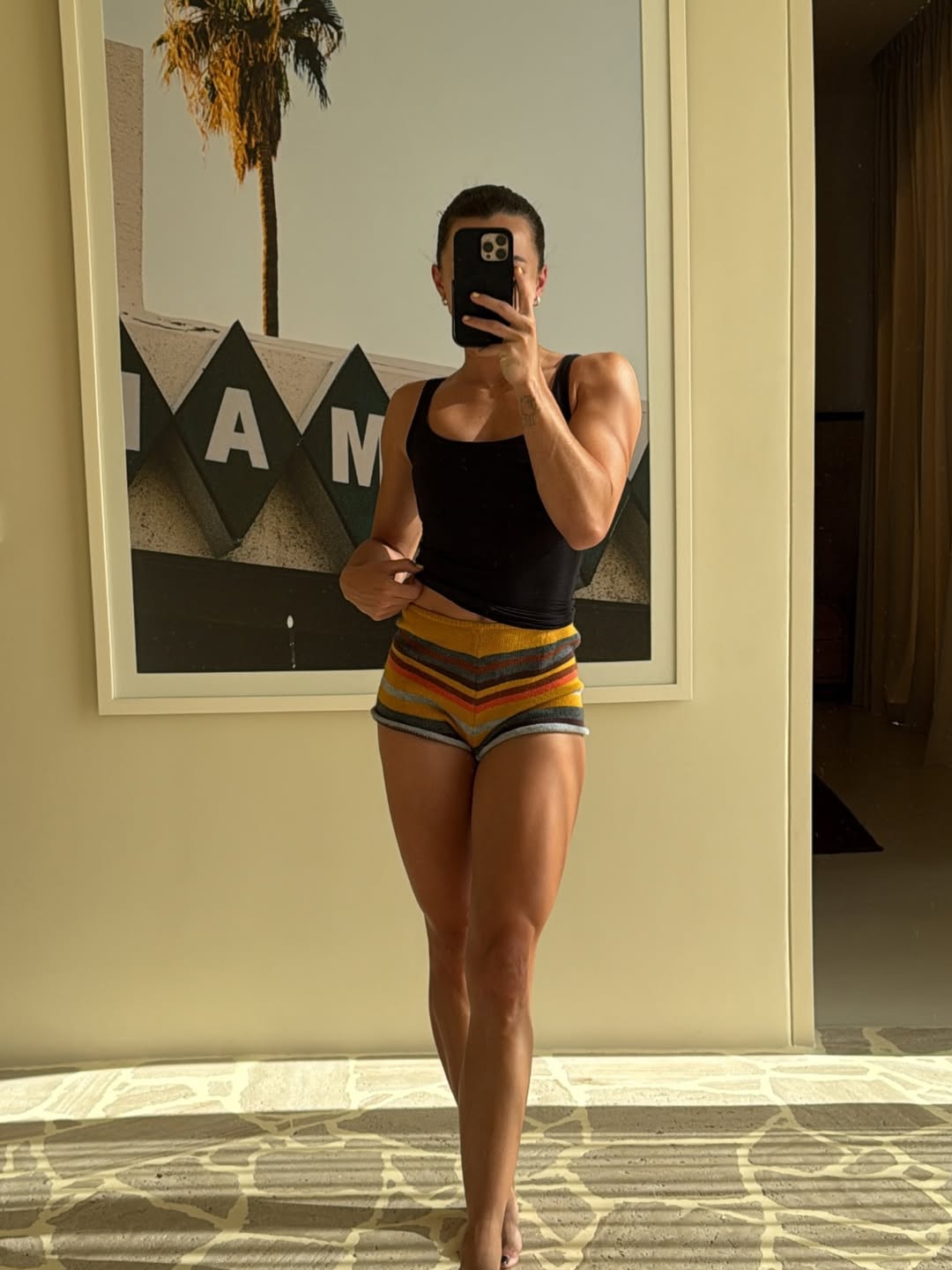 Person taking a mirror selfie with a PROPS iphone case  wearing a black tank top and colorful shorts in a room with a framed picture on the wall.