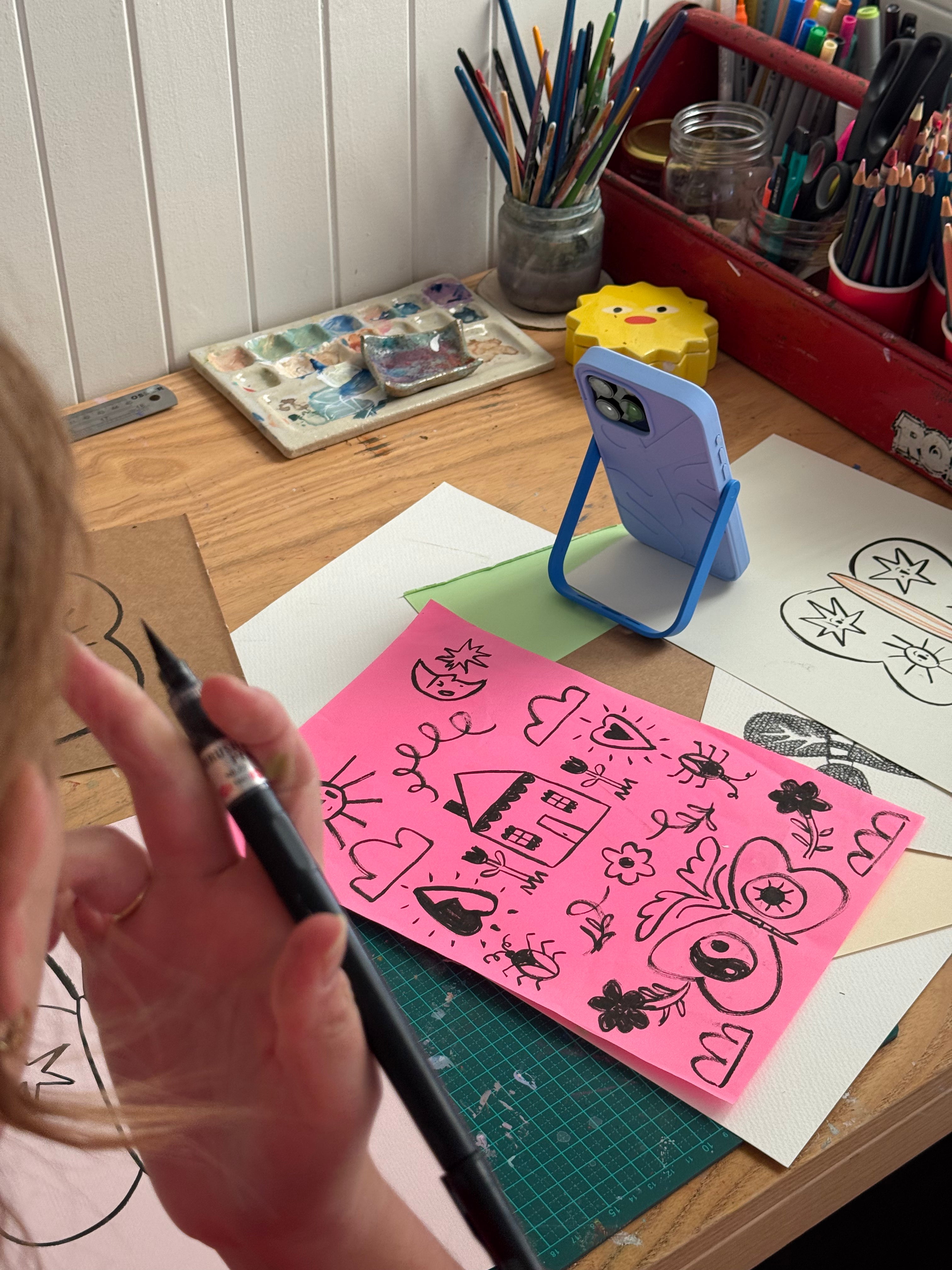 Illustrator drawing on pink paper, filming content on Props phone case surrounded by art supplies on a desk.